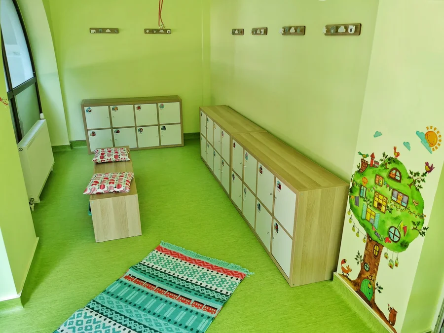 Vestiarul grădiniței: dulăpioare cu poze, o bancă, un covor și copacul pictat pe perete
